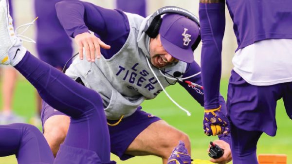 LSU Tigers football player in a purple cap and gray sweatshirt bends low during a training drill on the field.