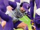 LSU Tigers football player in a purple cap and gray sweatshirt bends low during a training drill on the field.