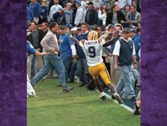 American football player in a white and yellow uniform with number 9 celebrates near the sideline as fans and bystanders crowd the field.