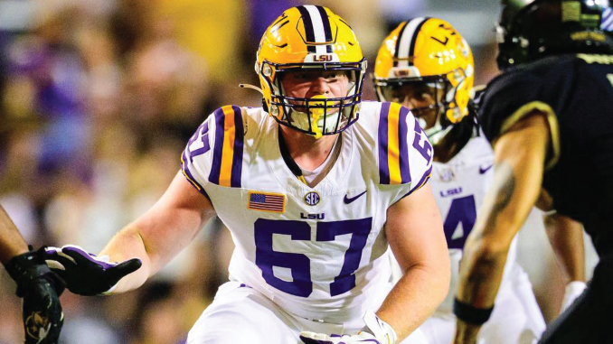LSU football player wearing number 67 in a white jersey with a gold helmet on the field during a game, reaching forward in action.