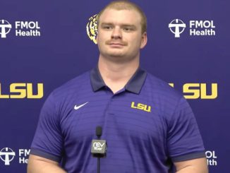 LSU athlete in a purple polo speaks at a press conference with a microphone in front and FMOL Health/LSU backdrop.