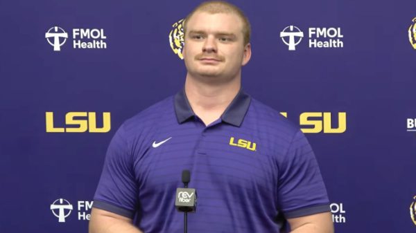 LSU athlete in a purple polo speaks at a press conference with a microphone in front and FMOL Health/LSU backdrop.