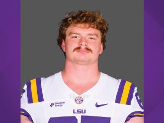 Portrait of a male LSU Tigers football player in a white jersey with purple and gold accents, facing the camera against a gray backdrop