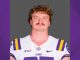 Portrait of a male LSU Tigers football player in a white jersey with purple and gold accents, facing the camera against a gray backdrop