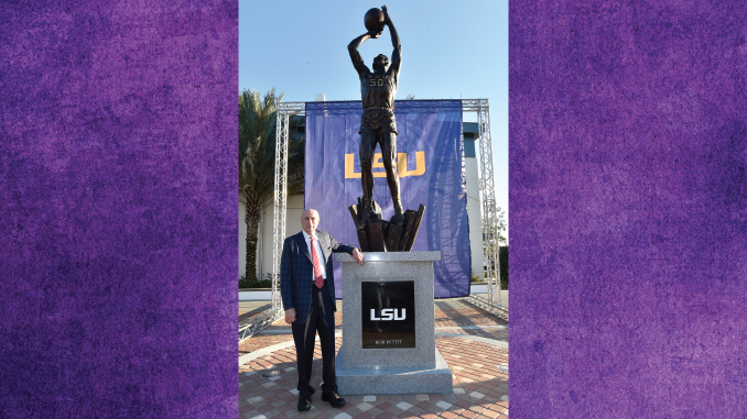 Statue of an athlete holding a ball overhead on a pedestal, with a man in a suit posing beside it and an LSU backdrop behind.