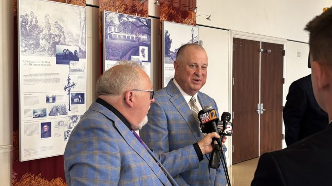 Man in a blue plaid suit being interviewed by reporters holding microphones in a hallway with historical exhibit panels on the wall behind him.