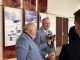 Man in a blue plaid suit being interviewed by reporters holding microphones in a hallway with historical exhibit panels on the wall behind him.