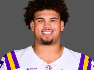 Portrait of LSU football player smiling, wearing a white jersey with purple and gold shoulders against a neutral gray background.