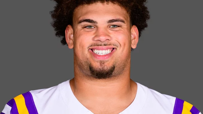 Portrait of LSU football player smiling, wearing a white jersey with purple and gold shoulders against a neutral gray background.