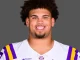 Portrait of LSU football player smiling, wearing a white jersey with purple and gold shoulders against a neutral gray background.