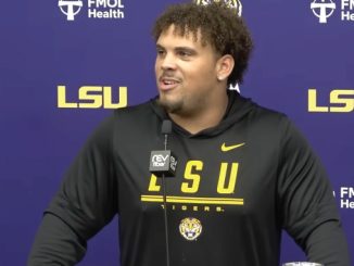 Young man with curly hair wearing a black LSU hoodie speaks at a press conference, microphone in front, purple backdrop with LSU and sponsor logos behind him.