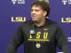 Young man with curly hair wearing a black LSU hoodie speaks at a press conference, microphone in front, purple backdrop with LSU and sponsor logos behind him.