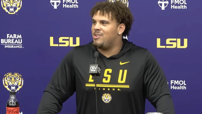 Young man with curly hair wearing a black LSU hoodie speaks at a press conference, microphone in front, purple backdrop with LSU and sponsor logos behind him.