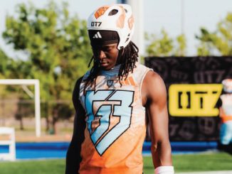 Football player in an orange sleeveless jersey and white helmet stands on a practice field, looking down.