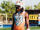 Football player in an orange sleeveless jersey and white helmet stands on a practice field, looking down.