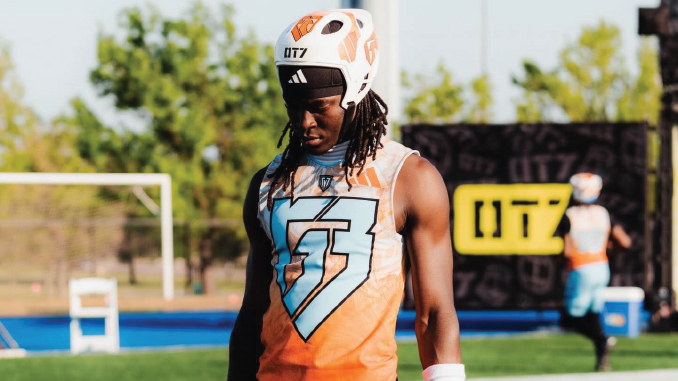 Football player in an orange sleeveless jersey and white helmet stands on a practice field, looking down.