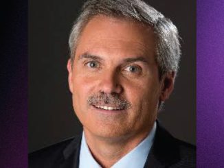 Head-and-shoulders portrait of a middle-aged man with gray hair and mustache, wearing a dark blazer and light-blue shirt against a dark background.