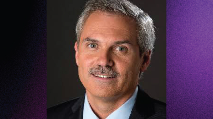 Head-and-shoulders portrait of a middle-aged man with gray hair and mustache, wearing a dark blazer and light-blue shirt against a dark background.