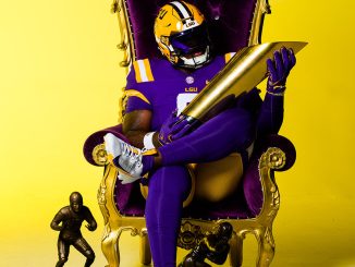 LSU football player in a purple and gold uniform sits in an ornate gold throne, holding a large gold trophy with three smaller trophies nearby against a yellow backdrop.