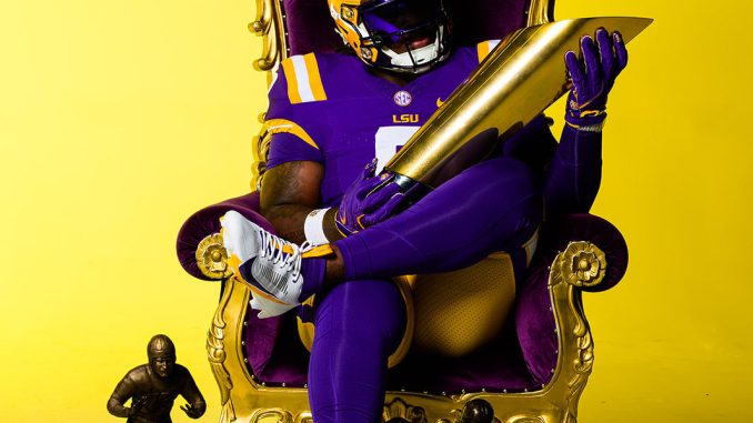 LSU football player in a purple and gold uniform sits in an ornate gold throne, holding a large gold trophy with three smaller trophies nearby against a yellow backdrop.