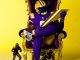 LSU football player in a purple and gold uniform sits in an ornate gold throne, holding a large gold trophy with three smaller trophies nearby against a yellow backdrop.