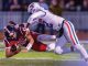 Red-uniform football player is tackled by a white-uniform defender on a turf field midair during a game, with the ball carrier clutching the ball.]","A red team ball carrier is brought down by a white team defender as they collide on the sideline turf.