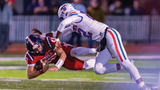 Red-uniform football player is tackled by a white-uniform defender on a turf field midair during a game, with the ball carrier clutching the ball.]","A red team ball carrier is brought down by a white team defender as they collide on the sideline turf.