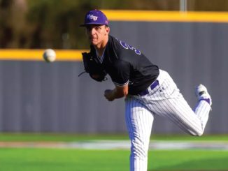 Baseball pitcher in a black jersey and purple cap unleashes a pitch on a sunny field, ball just leaving his hand.