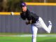 Baseball pitcher in a black jersey and purple cap unleashes a pitch on a sunny field, ball just leaving his hand.