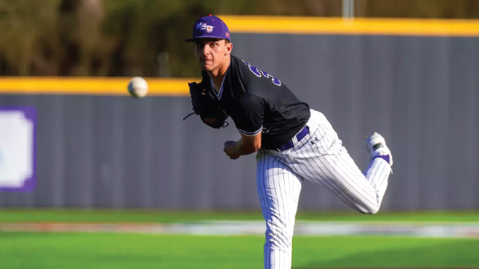 Baseball pitcher in a black jersey and purple cap unleashes a pitch on a sunny field, ball just leaving his hand.