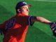 Baseball pitcher in a red jersey and blue cap mid-throw on a green field, glove extended outward.