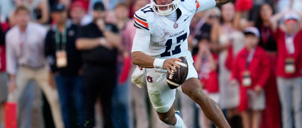 Auburn football player wearing number 11 in a white jersey runs with the ball on the field, crowd in the blurred stands behind him.