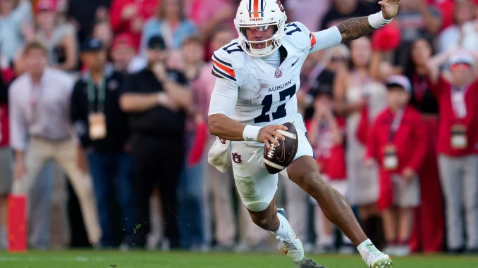 Auburn football player wearing number 11 in a white jersey runs with the ball on the field, crowd in the blurred stands behind him.