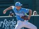Baseball pitcher in a light blue Carolina uniform winding up to throw a pitch on the mound with a brown glove, a sign and spectators in the background.