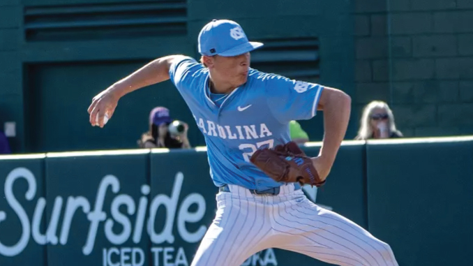 Baseball pitcher in a light blue Carolina uniform winding up to throw a pitch on the mound with a brown glove, a sign and spectators in the background.