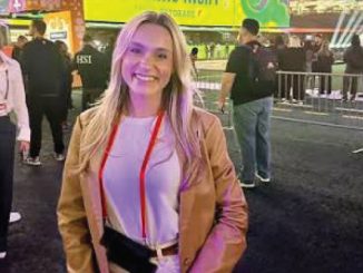 Smiling woman in a tan jacket and white top at a crowded expo, colorful booths behind her.