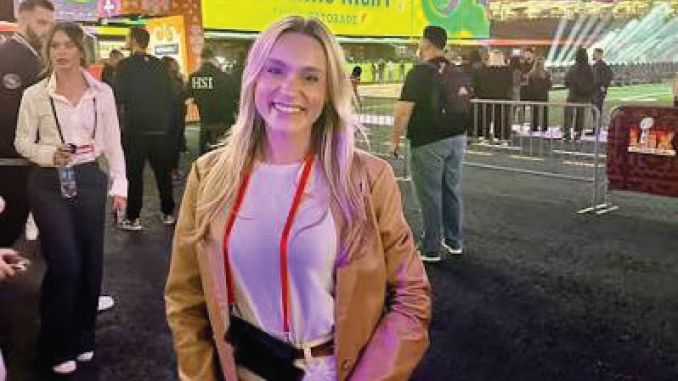 Smiling woman in a tan jacket and white top at a crowded expo, colorful booths behind her.