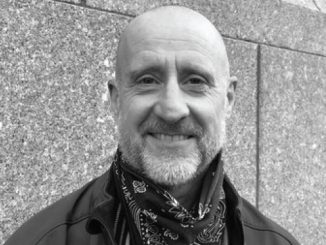Smiling bald man with a beard wearing a bandana scarf and jacket, standing against a stone wall.