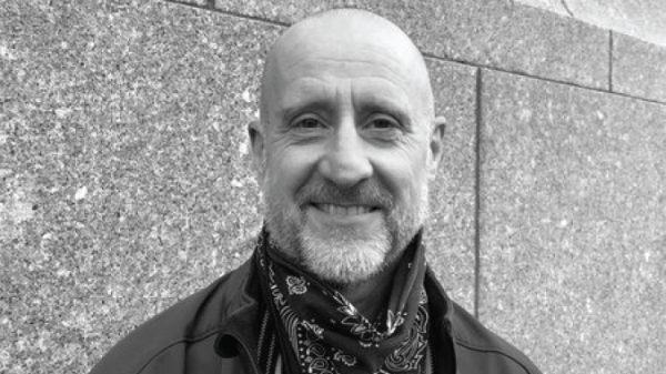 Smiling bald man with a beard wearing a bandana scarf and jacket, standing against a stone wall.