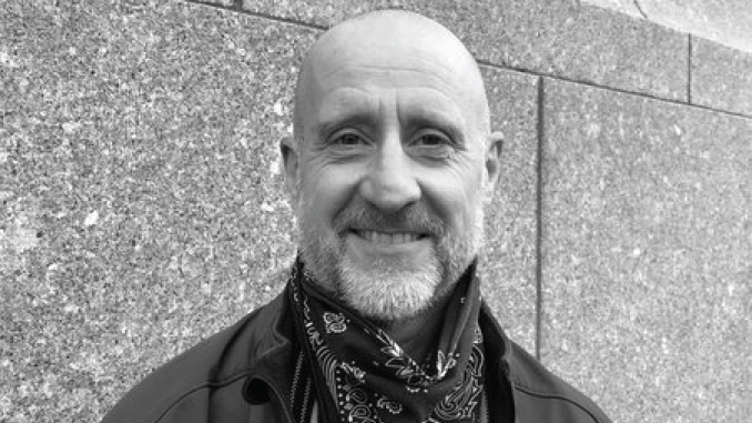 ChrisSmithVanityFair | TigerRag.com Smiling bald man with a beard wearing a bandana scarf and jacket, standing against a stone wall.