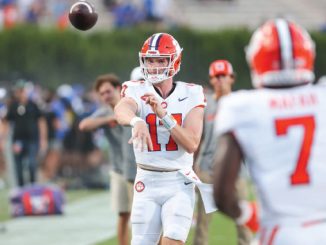 Quarterback in white uniform with orange helmet throws a pass on the field while a teammate wearing #7 stands nearby.