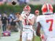 Quarterback in white uniform with orange helmet throws a pass on the field while a teammate wearing #7 stands nearby.
