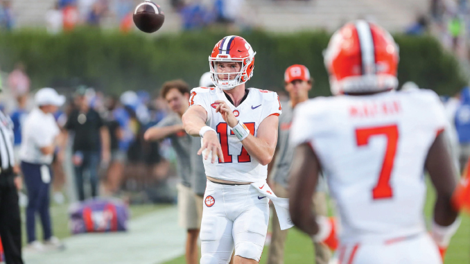 Quarterback in white uniform with orange helmet throws a pass on the field while a teammate wearing #7 stands nearby.