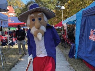 Mascot character with oversized beige beard and hat, wearing a blue coat and red pants, walking along an outdoor fair path with vendor tents in the background.