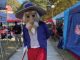 Mascot character with oversized beige beard and hat, wearing a blue coat and red pants, walking along an outdoor fair path with vendor tents in the background.