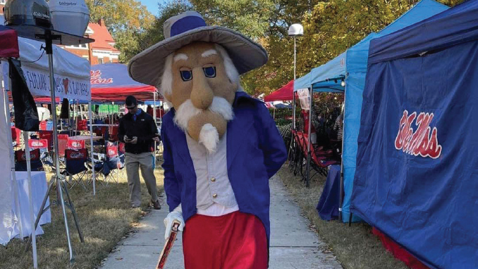 ColReb_OleMissAthletics | TigerRag.com Mascot character with oversized beige beard and hat, wearing a blue coat and red pants, walking along an outdoor fair path with vendor tents in the background.