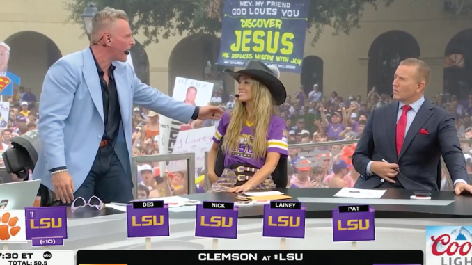 TV sports talk set with four anchors; a man in a light-blue blazer speaks to a woman in an LSU purple jersey and cowboy hat, as a suited host watches, with a large crowd and a 'Discover Jesus' sign in the background.