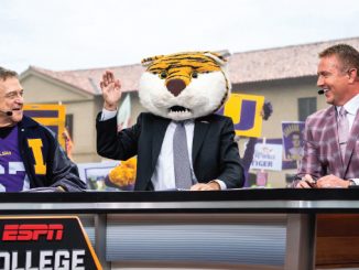 Tiger mascot in a suit waves from a broadcast desk between two hosts during a college sports segment on ESPN College set with a cheering crowd in the background, one host wearing a purple LSU jacket on the left.