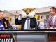 Tiger mascot in a suit waves from a broadcast desk between two hosts during a college sports segment on ESPN College set with a cheering crowd in the background, one host wearing a purple LSU jacket on the left.