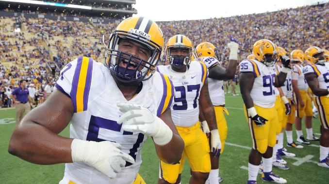 LSU football player in white jersey and yellow pants (#7) celebrating on the field with teammates; crowded stadium in background.